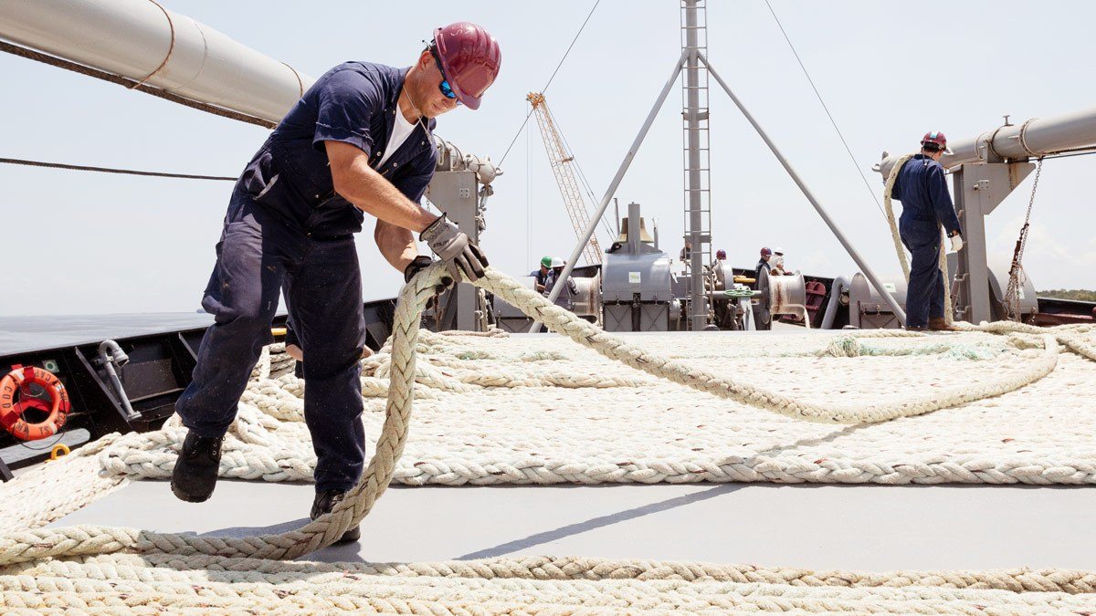 A mariner in a maroon hardhat and blue coveralls lays rope on the deck of a ship