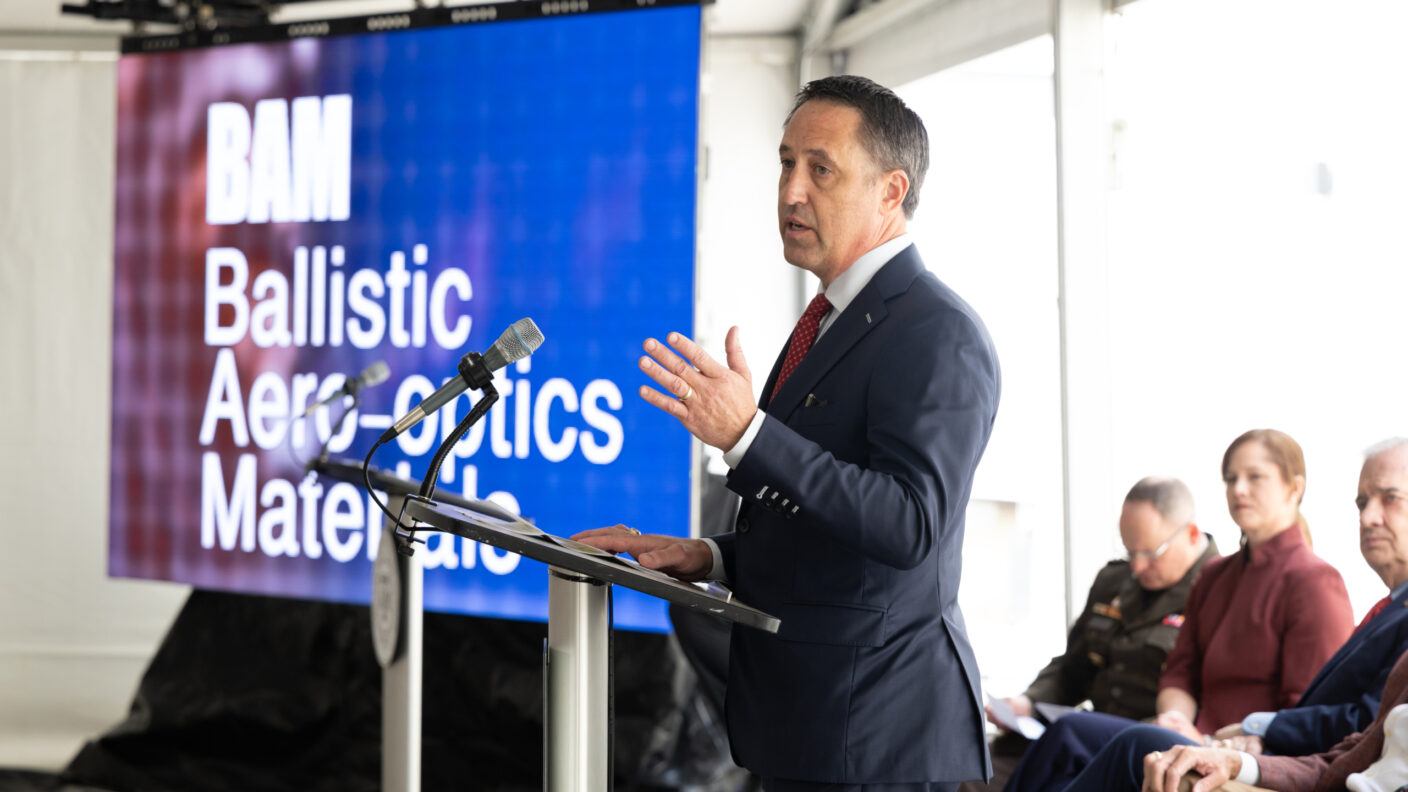 A man speaks at a lectern with people seated behind him at an event in front of an audience.
