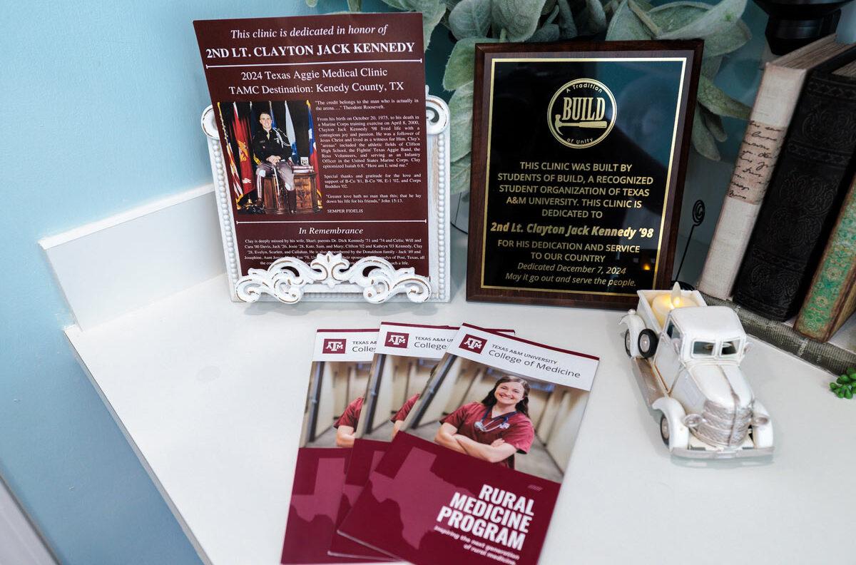 A plaque and small poster explain the Kenedy County Medical Clinic's dedication to 2nd. Lt. Clayton Jack Kennedy alongside brochures for the Texas A&M Rural Medical Program.