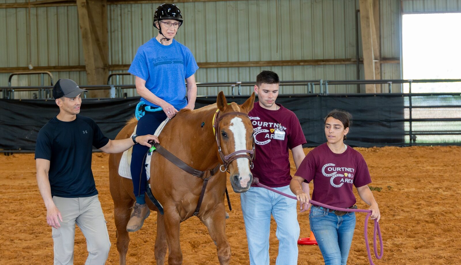 A photo of a person riding a horse in an arena with help from three people surrounding the horse on the ground.