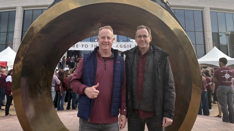 A photo of two men standing in front at the Aggie Ring Plaza in front of the Aggie Ring Statue.