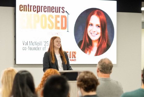 A photo of a woman to an audience at an event with a slide on a screen showing an image of herself in the background.