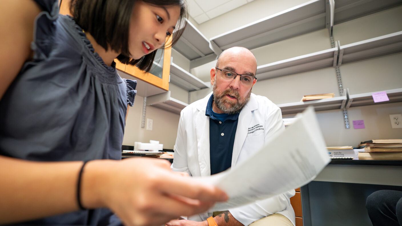 A female student holds a piece of paper in her hand while a professor in a white lab coat looks on