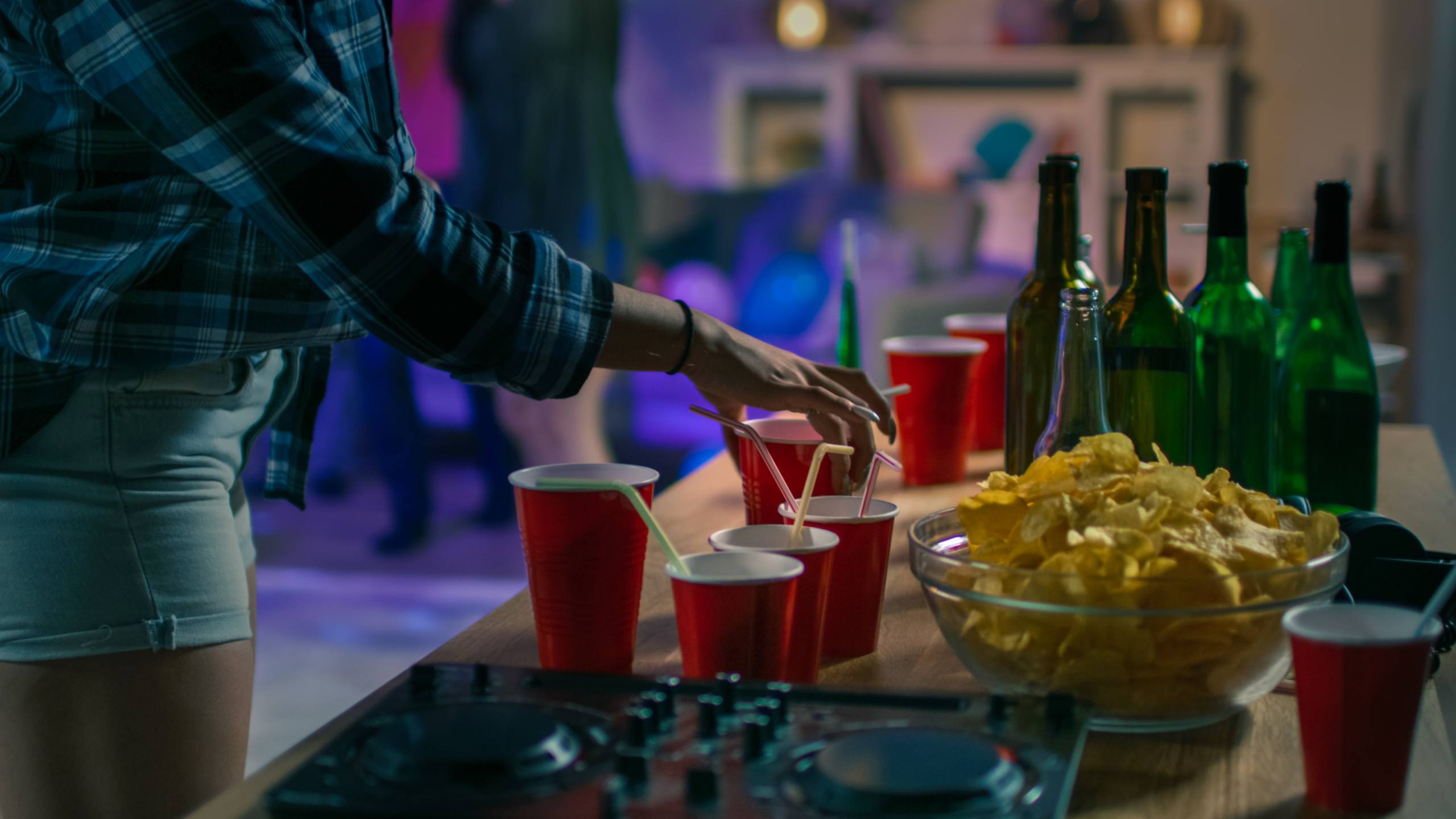 A photo of a table at a p arty with drink cups, bottles and chips, with a person reason for a drink cup.