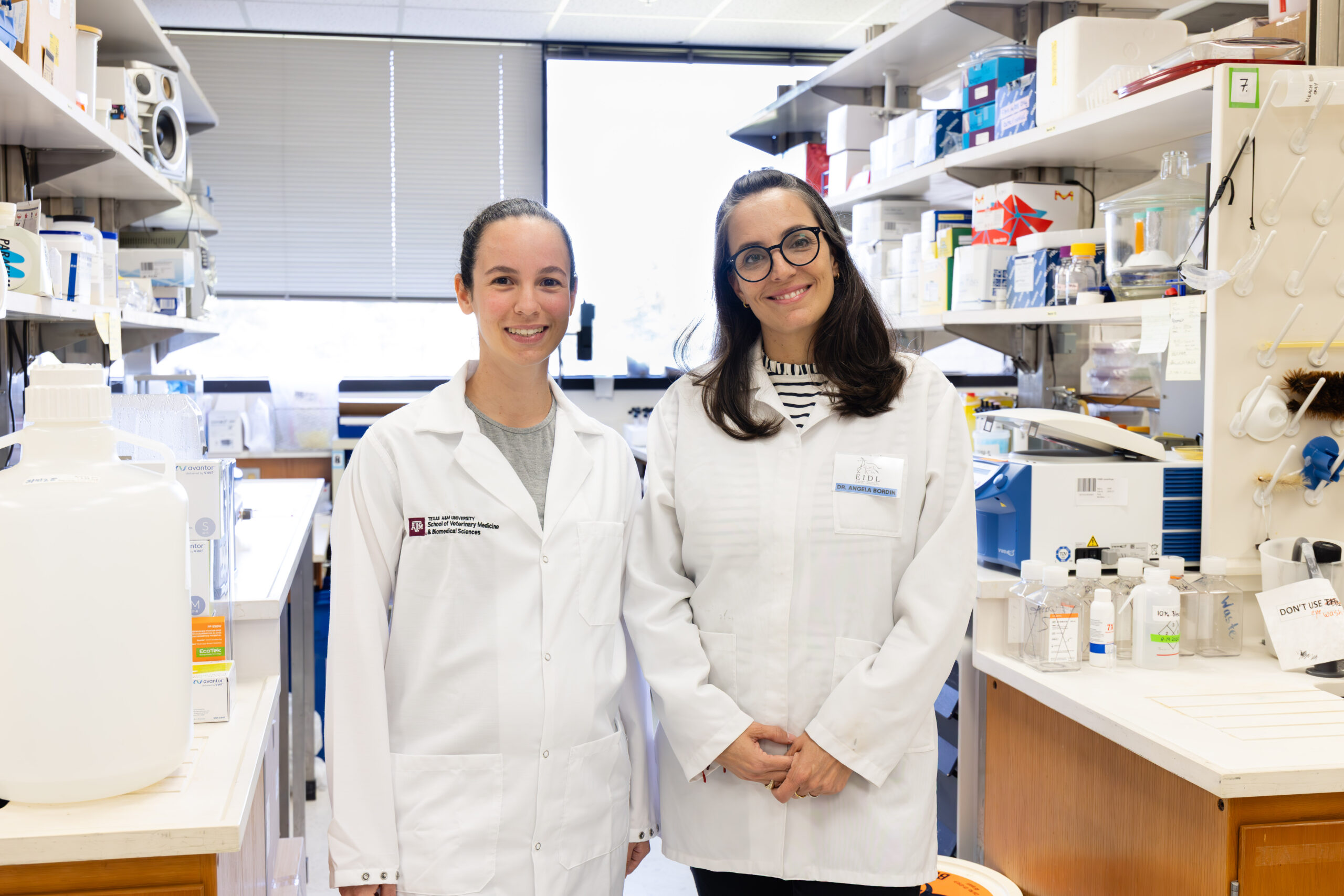 A photo of two women in lab coats posing for a photo among shelves in a research laboratory.