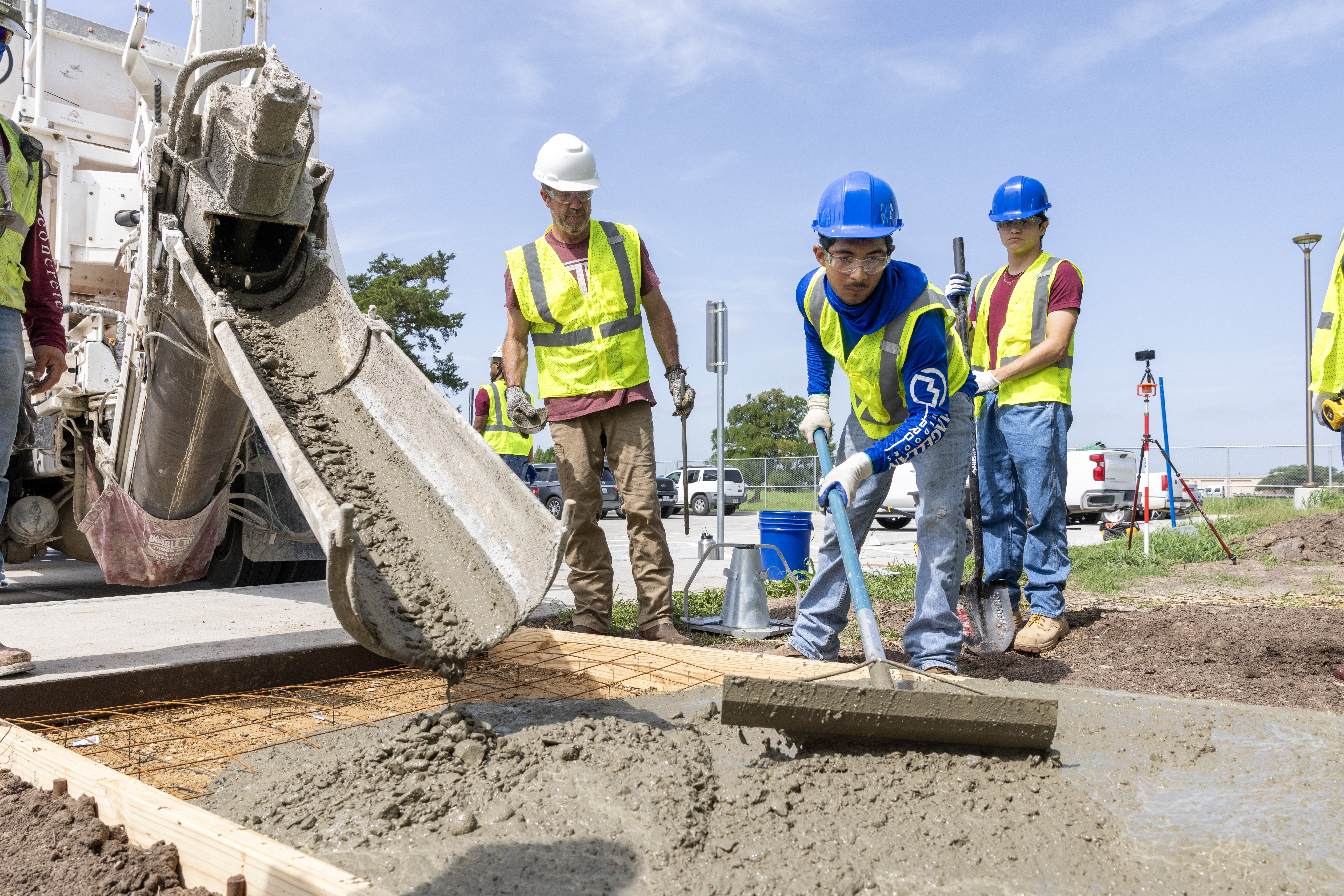 A student in a hard hat and yellow high-visibility vest spreads wet concrete from a mixer truck while being supervised by an instructor. Another student in safety gear observes.