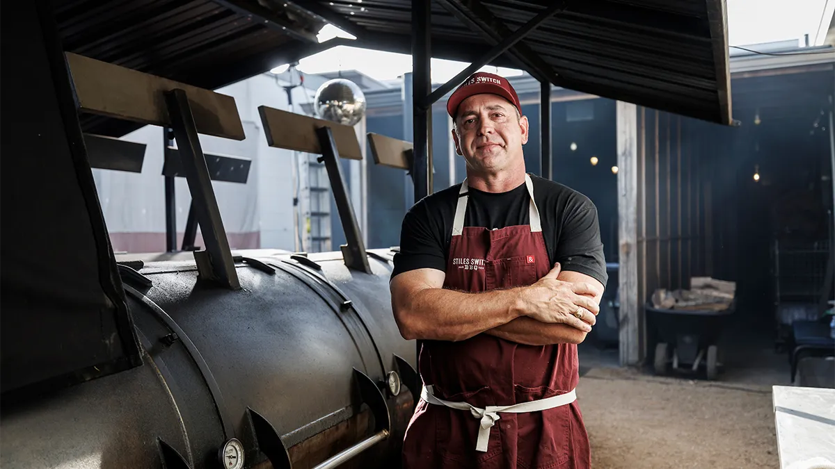 Portrait of Shane Styles of wearing a maroon apron and his Aggie Ring in front of a BBQ pit