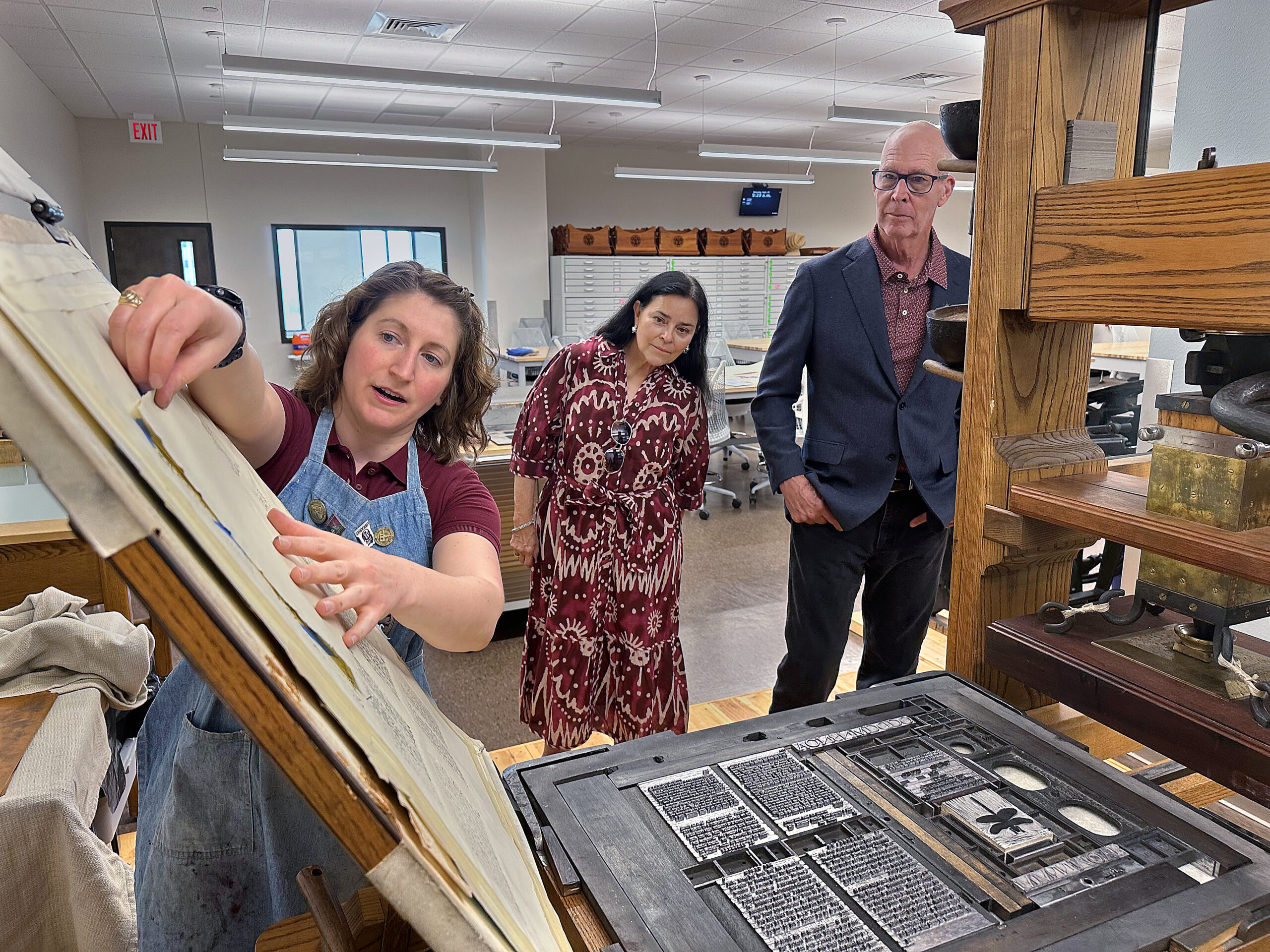 A photo of a librarian showing a historic press to a man and a woman.
