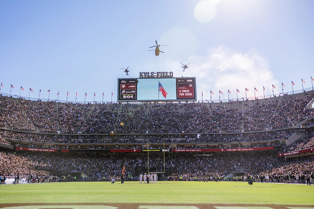 Military helicopters fly over the Kyle Field score board that features a waving American flag.