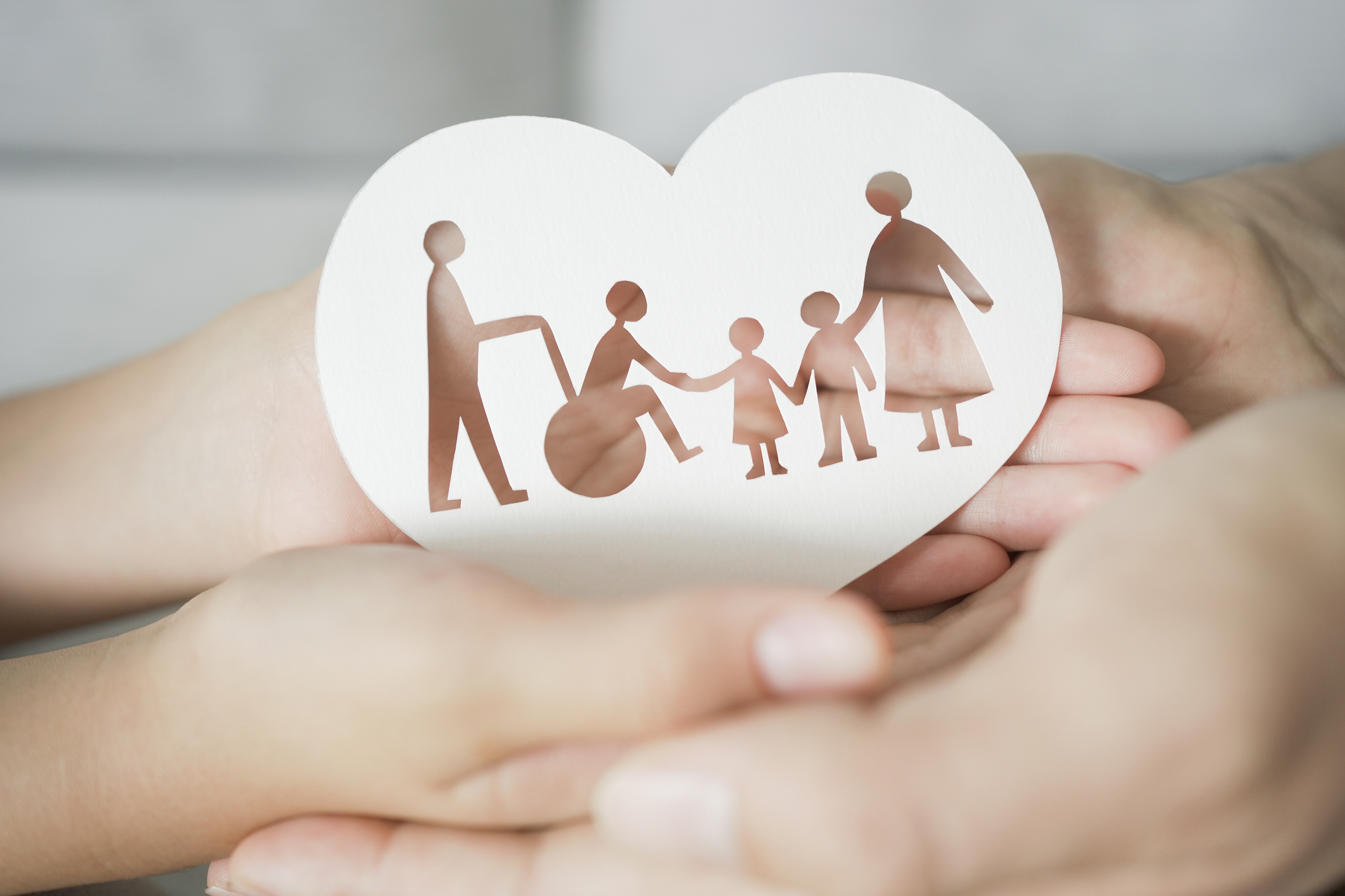 hands holding a heart-shaped paper cutout of a woman in a wheelchair with people of other generations around her